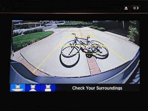 Rearview camera on a center screen showing a bicycle in the driveway behind the car, with yellow guide lines and the message Check Your Surroundings.