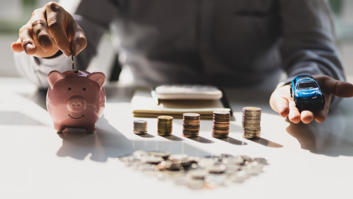 A man holds a piggy bank and a toy car next to stacks of coins