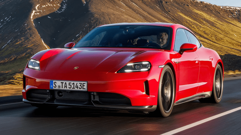 A red 2026 Porsche Taycan driving towards the camera in a left-side 3/4 view on a paved road with red sandstone hills behind it.